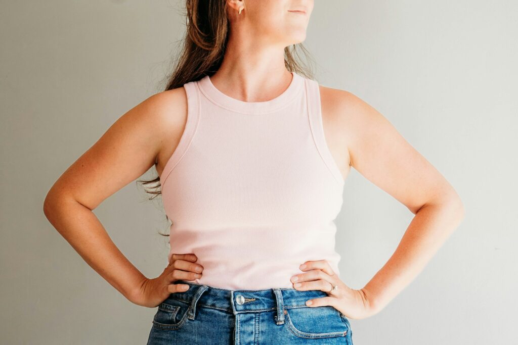 Person wearing a light pink sleeveless top and jeans stands with hands on hips, facing left.